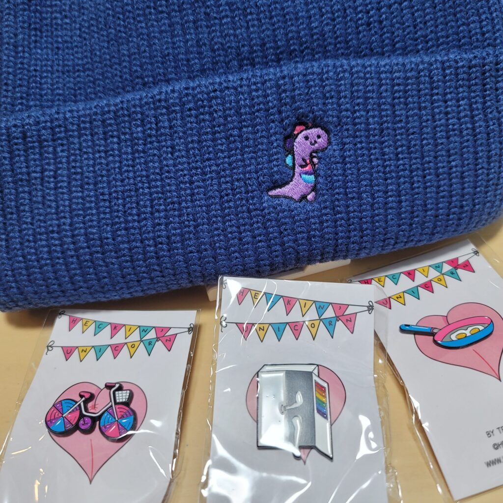 画面奥にバイセクシュアルフラッグカラーの恐竜の刺繍が入った青いニット帽、手前左からバイセクシュアルフラッグカラーの自転車のピンズ、レインボーカラーが入ったクローゼットから出てくる人のピンズ、パンセクシュアルフラッグカラーのフライパンのピンズ