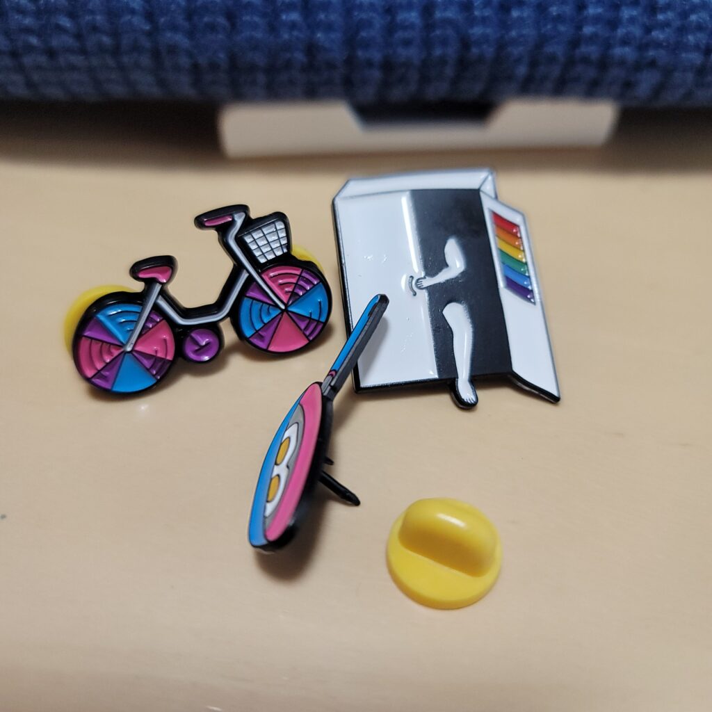 手前左からバイセクシュアルフラッグカラーの自転車のピンズ、レインボーカラーが入ったクローゼットから出てくる人のピンズ、パンセクシュアルフラッグカラーのフライパンのピンズ。フライパンのピンズは留め具を外している